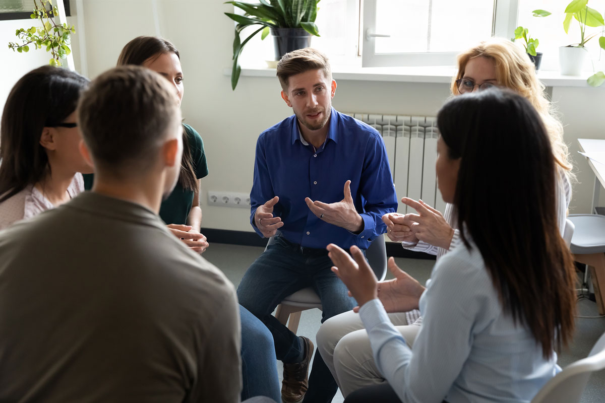 In a bright room overlooking NYC, a diverse group of people is seated in a circle engaged in discussion. One person in a blue shirt speaks while others listen attentively, much like an impromptu therapy session. Potted plants adorn the windowsill in the background.