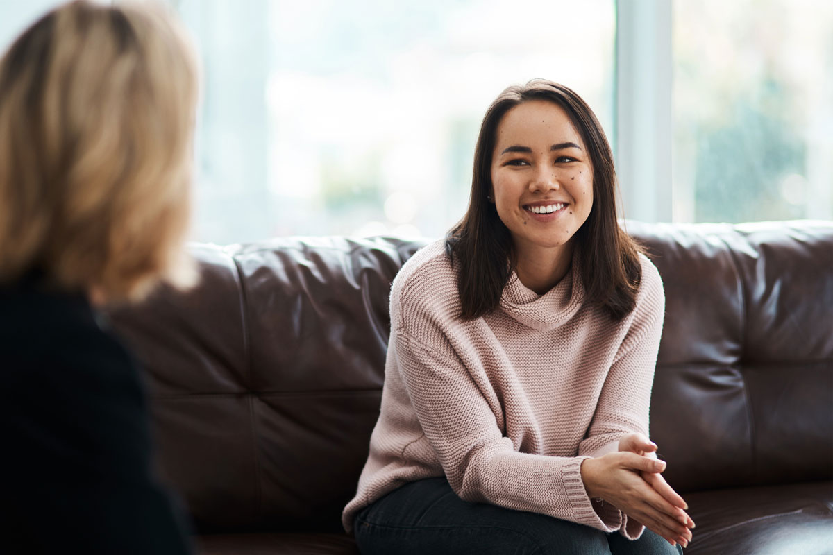 A woman with long dark hair, wearing a pink sweater, sits on a brown leather couch in a bright NYC room, smiling and gesturing as if discussing therapy. Another person with blonde hair is blurred in the foreground near large windows.