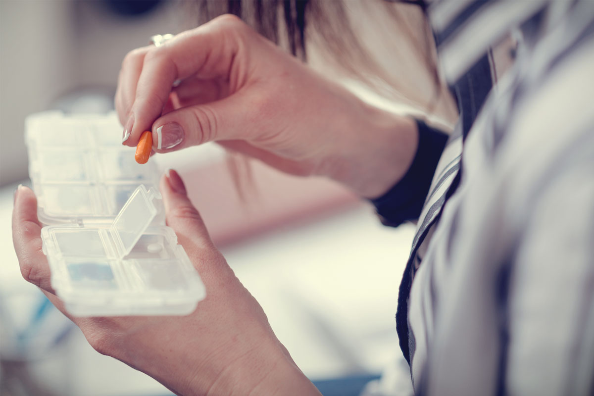 A person is holding an orange pill and a pill organizer, suggesting they are sorting their medication as part of their therapy routine. The backdrop is a blurred NYC skyline, directing focus on the hands deftly arranging the organizer's open compartments.