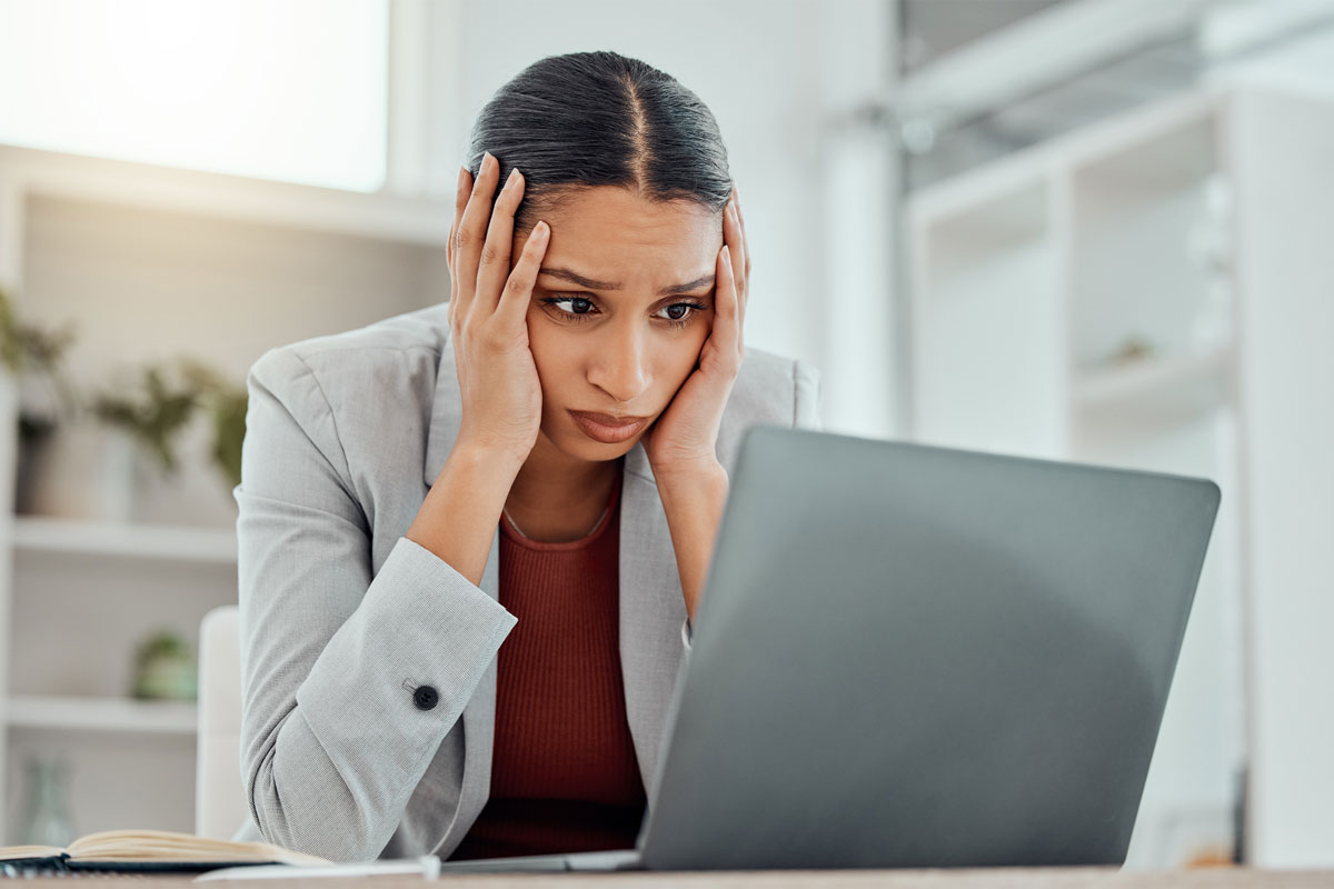 In a bright, modern NYC office, a woman in a blazer looks stressed, resting her head in her hands as she stares at a laptop screen. The scene conveys a sense of concern or frustration, perhaps hinting at the need for some much-deserved therapy.