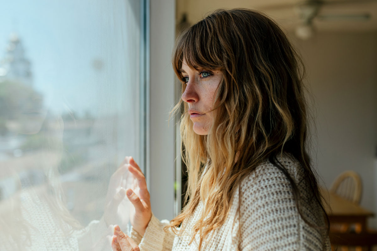 A woman with long hair gazes thoughtfully out a window, her hand touching the glass. She is indoors in NYC, wearing a knit sweater, with soft lighting illuminating her reflective expression. The background is slightly blurred, hinting at a cozy room that feels like therapy for the soul.