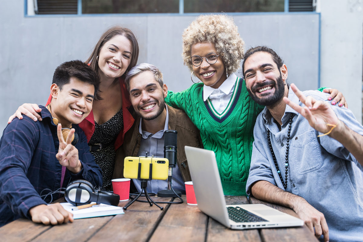 Around a wooden table in NYC, a diverse group of five people smiles and poses for a photo with a laptop, microphone, and smartphone stand. They appear cheerful and friendly, as if they've just wrapped up an uplifting therapy session; two are even making peace signs.