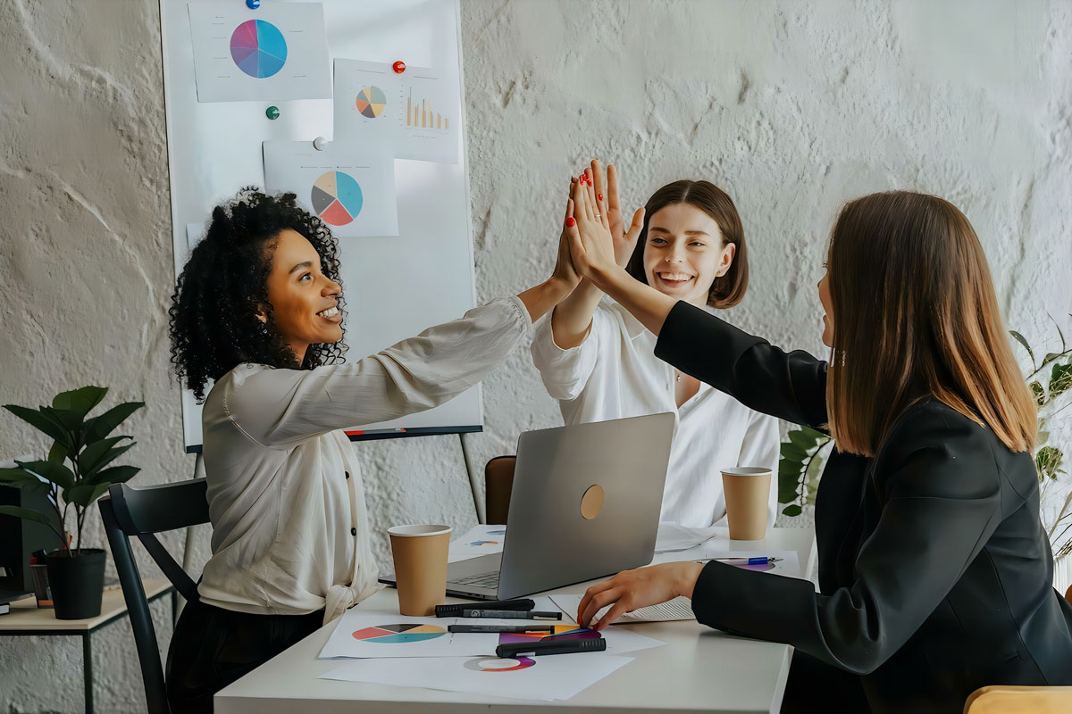 Three women sit around a table with charts, a laptop, and coffee cups. Two of them are high-fiving while smiling, suggesting teamwork or a successful collaboration in the midst of an NYC office. A whiteboard with graphs is in the background.