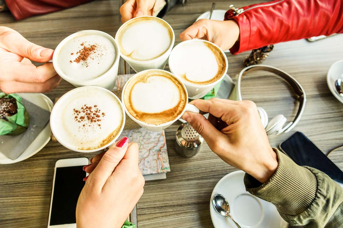 Five people clink coffee mugs over a modern table in NYC. Their frothy cappuccinos, some dusted with cocoa. On the table lie a smartphone, headphones, a map, and an empty saucer.