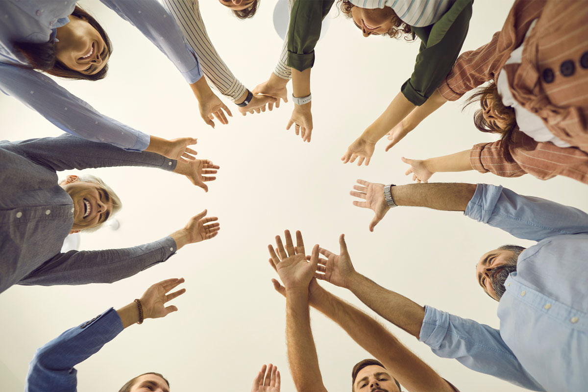 A diverse group of people stands in a circle amid the NYC skyline, reaching their hands towards the center and looking upwards. The photo, taken from below, captures a sense of unity and teamwork against a soft, neutral background.