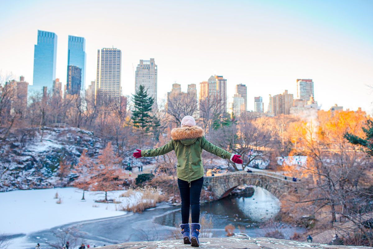 A person in a green coat stands with arms outstretched, facing the NYC skyline. Tall buildings and a small bridge hover over a partially frozen pond. The park's winter scene, where snow blankets the ground and trees, offers a quiet therapy for the soul.