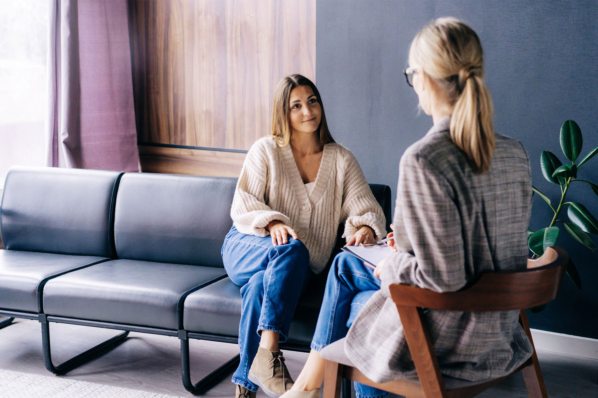 In a modern NYC office, two women sit in a serene setting. One is on a black couch, wearing a beige sweater and jeans, listening attentively. The other, in a plaid blazer and glasses, sits on a wooden chair with a clipboard, deep in therapy conversation. A plant adorns the background.