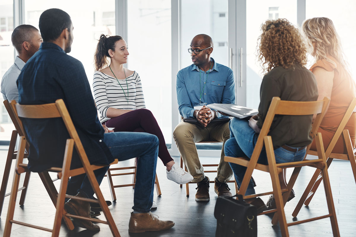 In a well-lit NYC office, five people—three men and two women—sit in a circle having a lively discussion. Engaged in the conversation, one man gestures as he speaks, embodying the essence of group therapy. All are attired in business casual wear.