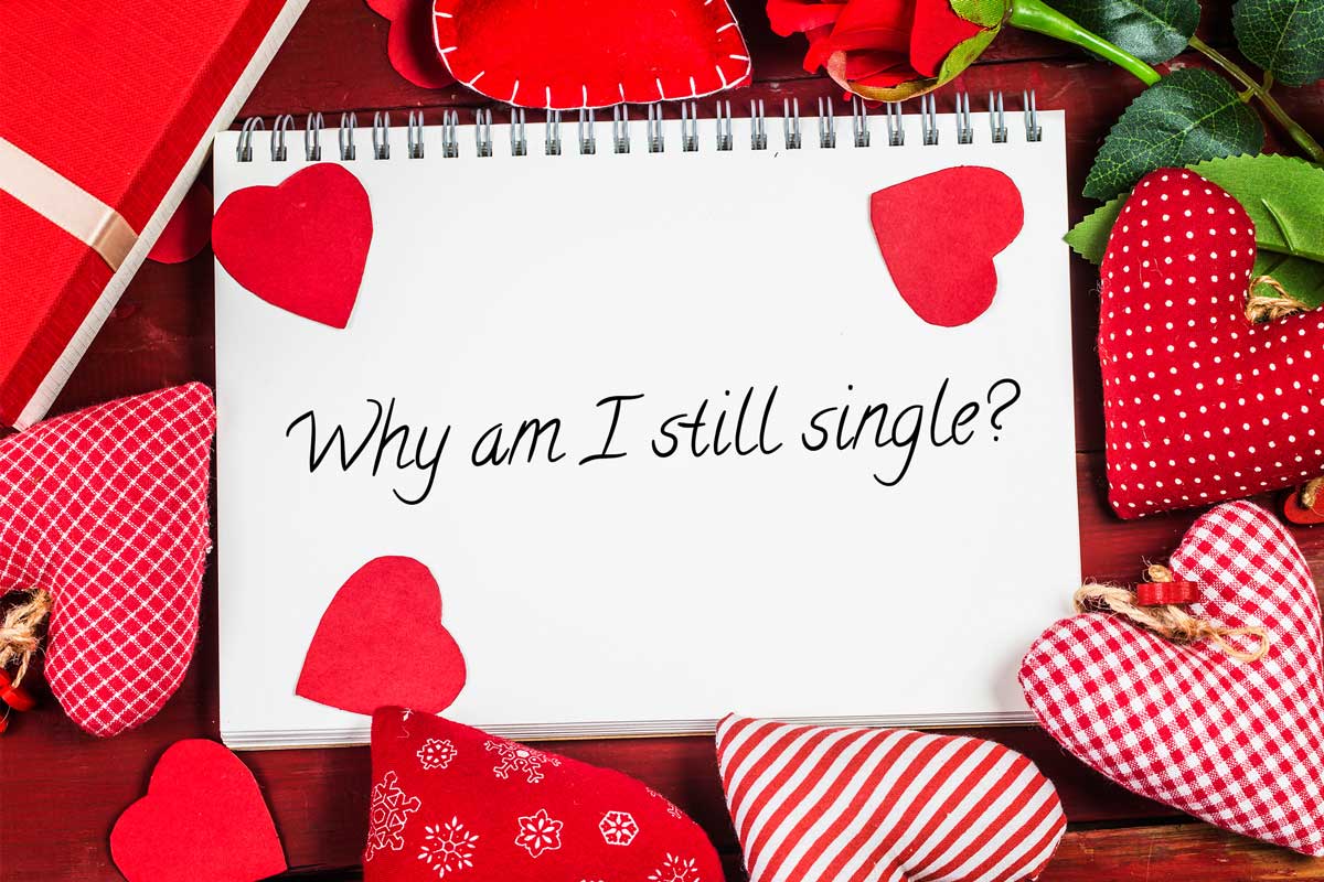 A notepad with "Why am I still single?" written in cursive is surrounded by red and checkered hearts, a red gift box, and a red rose on a wooden surface.