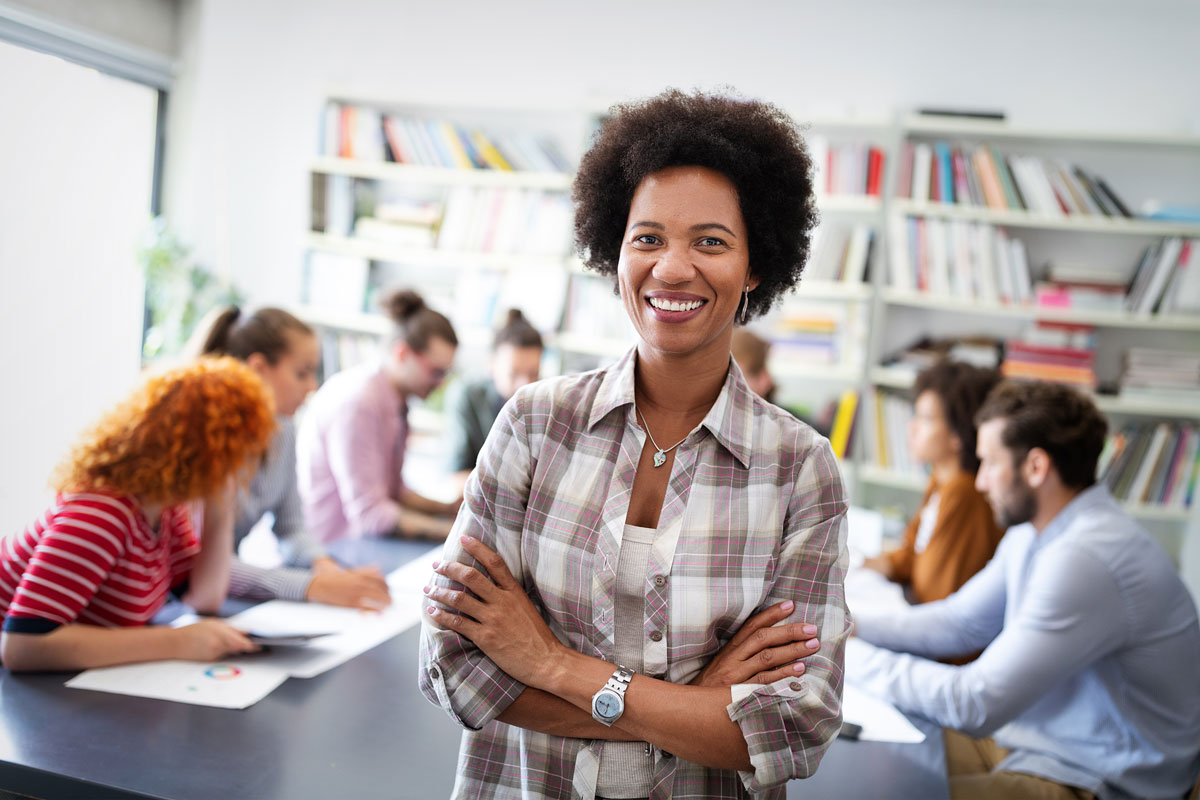 A confident woman with curly hair smiles and crosses her arms. In the background, a group engages in a lively discussion at a table in an NYC office filled with bookshelves, creating a space that feels both vibrant and therapeutic.