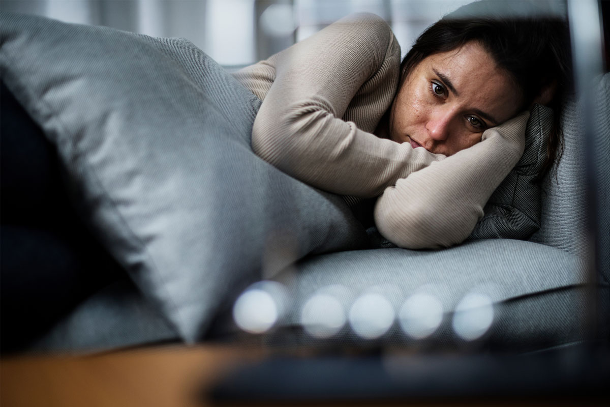 A person with long hair lies on a couch, resting their head on a gray pillow. They are wearing a beige sweater and appear pensive, as if lost in thoughts of NYC. The background is blurred, emphasizing their expression.