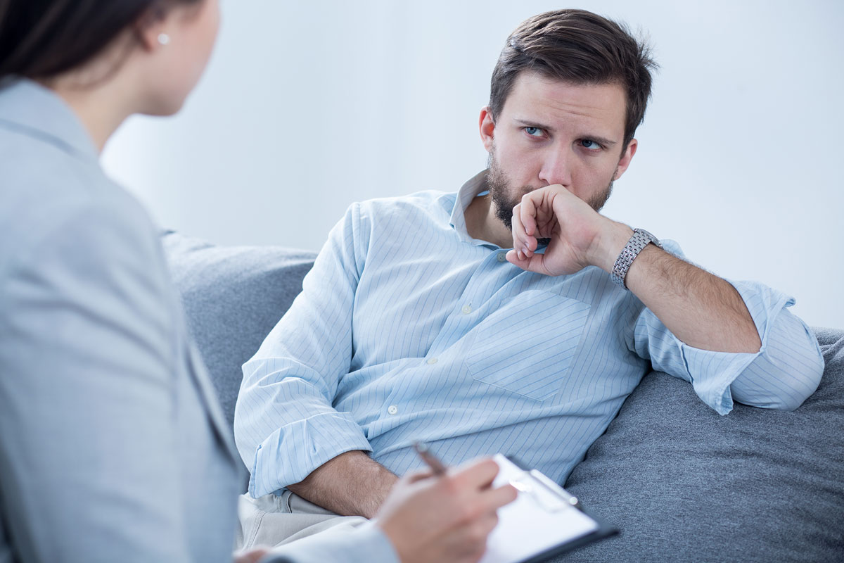 A man with a thoughtful expression sits on a couch, resting his chin on his hand. In this NYC setting, he listens to a woman holding a clipboard, partially visible, suggesting a therapy or counseling session.