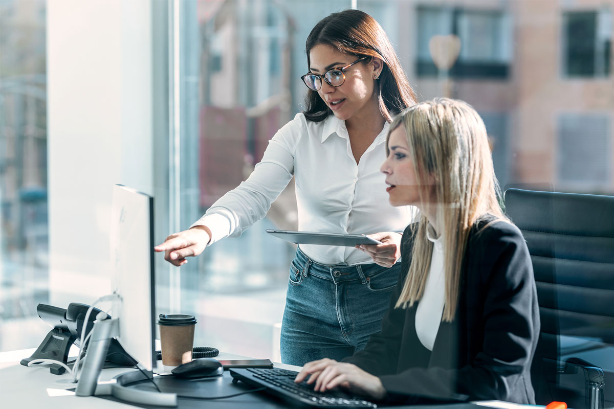 In a bustling NYC office, two women collaborate: one stands, pointing at a computer screen with her tablet like a seasoned therapist guiding through data, while the other types away. A coffee cup adds warmth as large windows frame the vibrant cityscape in the background.