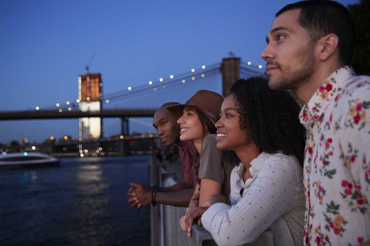 Four people are leaning on a railing, gazing across the water at dusk. The iconic Brooklyn Bridge and the NYC skyline form a stunning backdrop. They appear relaxed and content, as if the view itself is a gentle form of therapy.