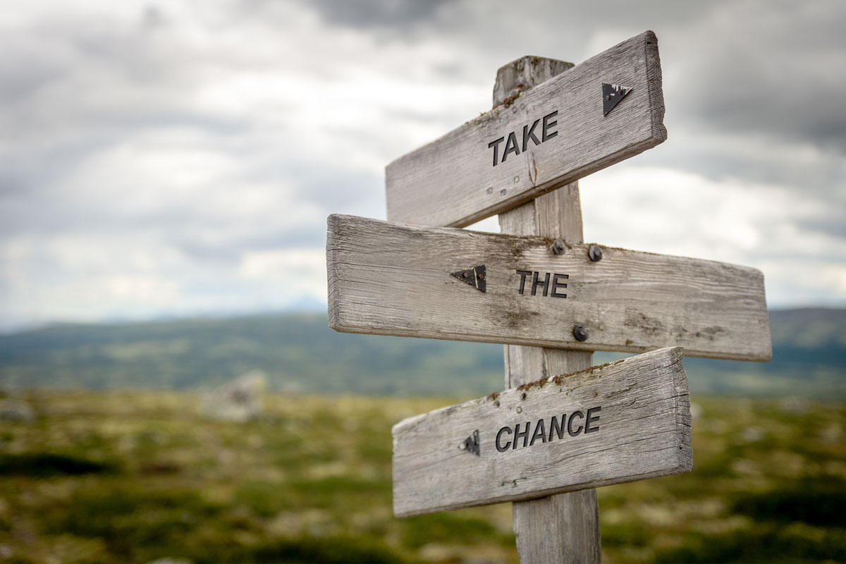 A wooden signpost with arrows pointing in three directions, featuring the words "TAKE," "THE," and "CHANCE" under a cloudy sky. The background reveals a blurred grassy landscape, reminiscent of a serene escape from the hustle and bustle of NYC, offering a touch of natural therapy.