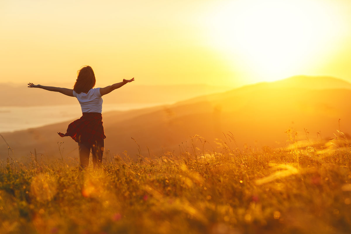 A person stands in a field with outstretched arms, embracing the therapeutic warmth of a golden sunset. The clear sky casts a serene glow over rolling hills and distant water, reminiscent of escaping NYC's hustle for nature's tranquil embrace.