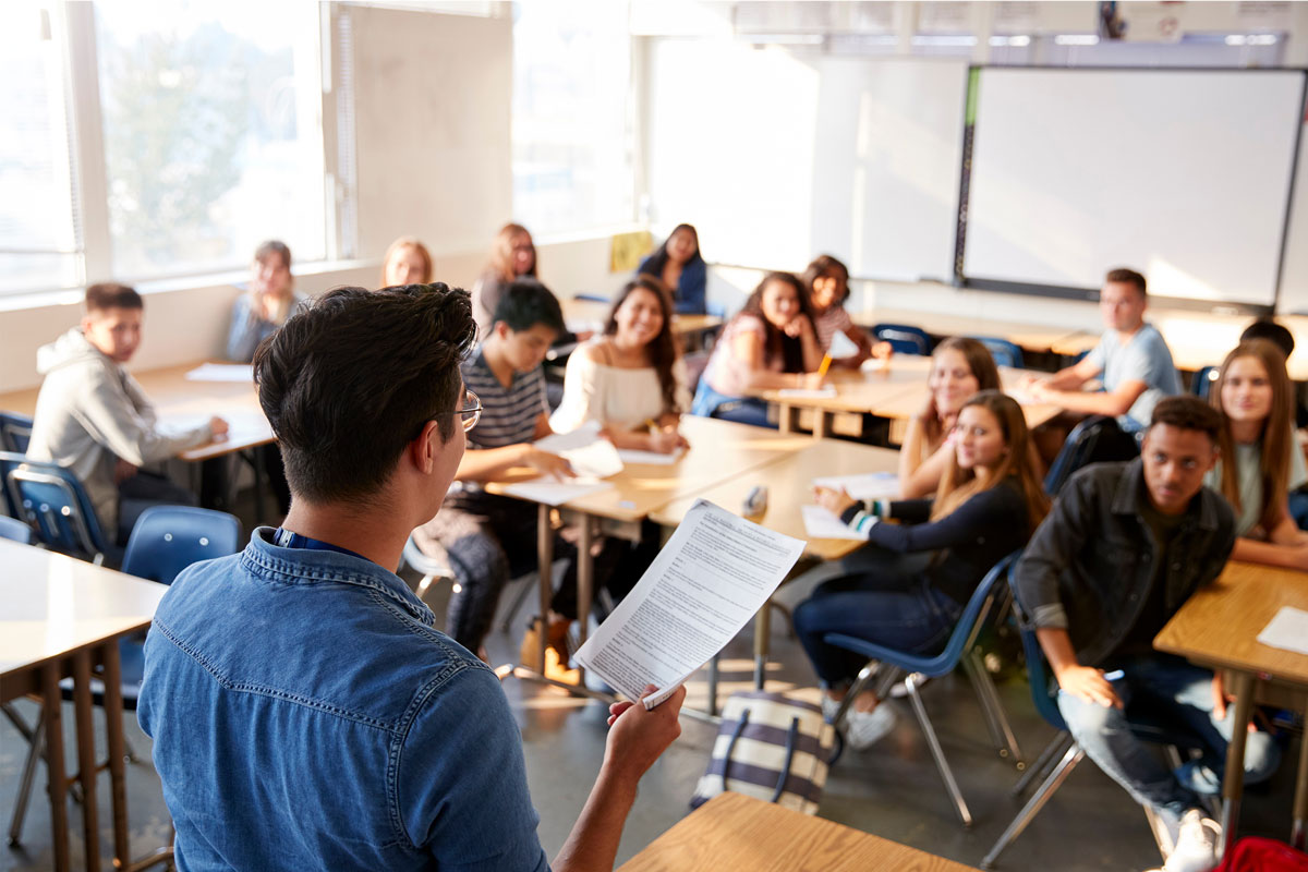 A teacher stands at the front of a classroom in NYC, holding and reading from a sheet of paper. Students are seated at desks, attentively listening. Sunlight filters through the windows, creating a warm, almost therapeutic atmosphere.