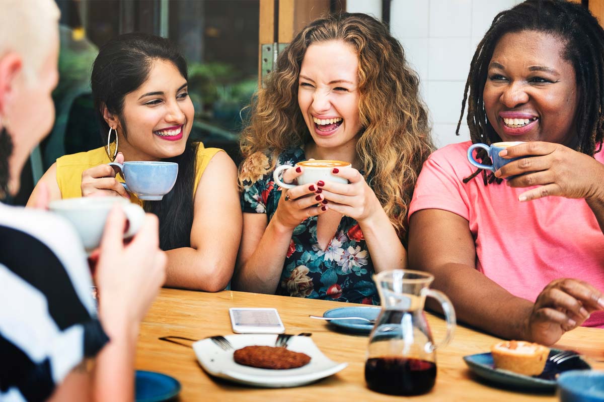 Four people sit at a wooden table in a cozy NYC café, smiling and holding cups of coffee. As they enjoy a casual conversation, with small plates of food before them, the warm atmosphere feels almost therapeutic.