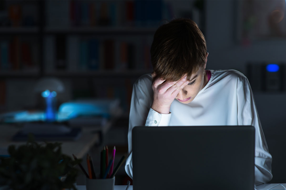 A person sits at a desk, leaning over a laptop with their hand on their forehead, appearing stressed or tired. The room resembles a cozy NYC nook, dimly lit with bookshelves blurred in the background and a lamp emitting a calming blue light—perhaps they could use some therapy.