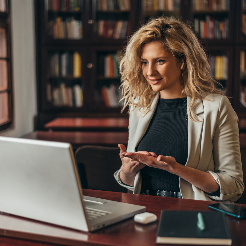In a cozy library setting, a woman with curly blonde hair sits at her desk in NYC, smiling and gesturing as if conducting an online therapy session. Bookshelves line the background, while a smartphone and notebook rest beside her laptop.