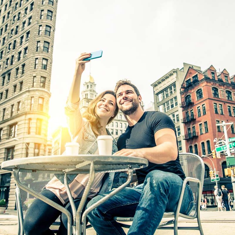 A smiling couple takes a selfie at an outdoor café table in NYC, with coffee cups before them. The sunny cityscape showcases historic buildings, including a flatiron building, offering a joyful backdrop that feels like therapy for the soul.