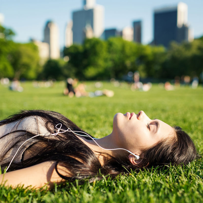 A woman with long dark hair lies on grass in a NYC park, eyes closed and listening to music with earphones. The city skyline and trees blur in the background. The sun casts a warm glow, creating a peaceful atmosphere that feels like nature's own therapy session.