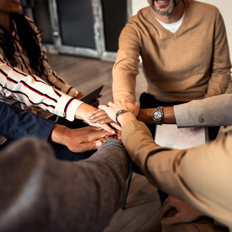 In a cozy NYC studio, a group gathered in a circle places their hands together in the center, symbolizing unity and teamwork. Dressed casually on the wooden floor, their connection exudes warmth and support—like an uplifting therapy session for the soul.