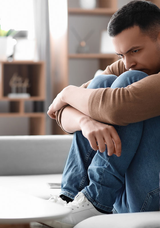 A man sits on a gray couch in a modern NYC living room, hugging his knees, looking pensive. He wears a brown sweater, blue jeans, and white sneakers. A laptop lies open on the table before him—a silent companion in his day of introspection. A plant adds life to the background.