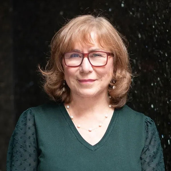 A woman with shoulder-length brown hair and glasses smiles at the camera. She is wearing a dark green top with sheer sleeves and a pearl necklace, exuding an air of NYC sophistication, as she stands against a dark, textured background.