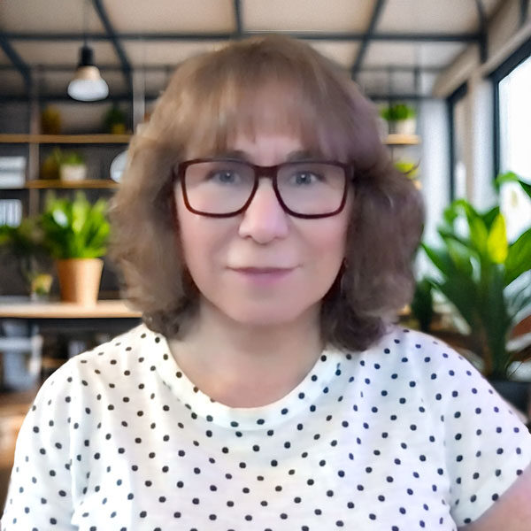 A person with shoulder-length hair and glasses is wearing a white polka dot shirt. In a cozy NYC setting, with shelves, hanging lights, and various potted plants in the background, it feels like a space for relaxation or therapy.