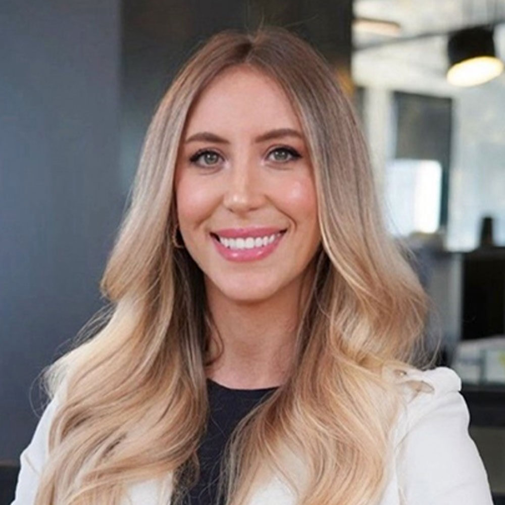 A woman with long blonde hair and a light-colored blazer smiles at the camera. She is in an NYC office setting, with blurred background elements such as lights and windows.