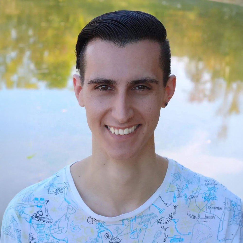 A smiling person with short dark hair and a doodle-covered white shirt stands in front of a reflective water surface, surrounded by lush greenery, evoking tranquility reminiscent of a serene therapy retreat far from the bustle of NYC.