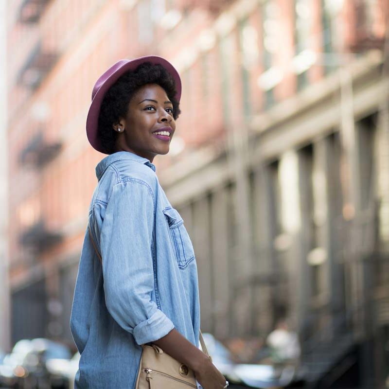 Woman with curly hair and pink hat, smiling as she strolls down an NYC street lined with tall buildings. She's wearing a denim shirt and carrying a beige bag, against the blurred urban backdrop—a perfect therapy for city lovers embracing the vibrant energy.